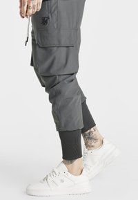 Pantalones cargo grises con un puño ajustado en el tobillo, que presentan bolsillos laterales y una tela texturizada. Combinados con zapatillas blancas.