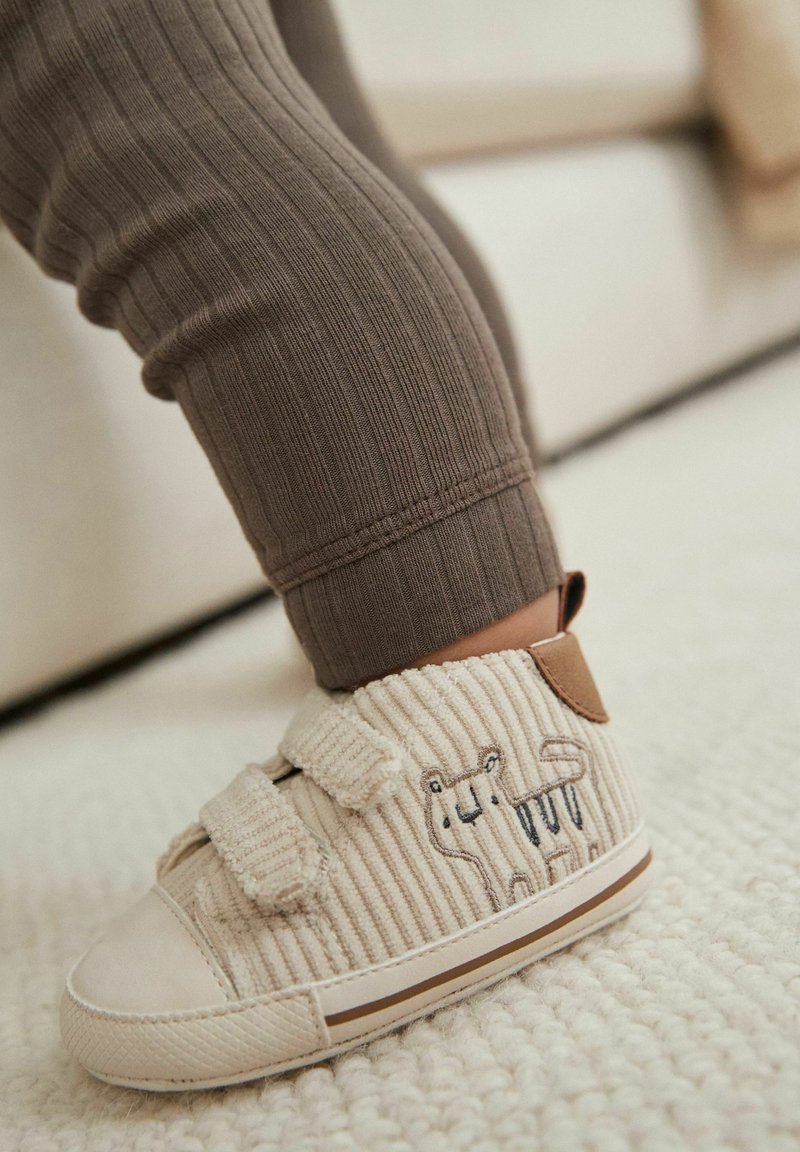 Pied d'enfant portant une chaussure beige en velours côtelé pour tout-petit avec broderie d'animal et bandes velcro, reposant sur un tapis crème texturé.