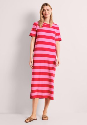 Kvinde iført en midi-kjole med røde og lyserøde vandrette striber, korte ærmer og brune flade sandaler, stående mod en ensfarvet baggrund.