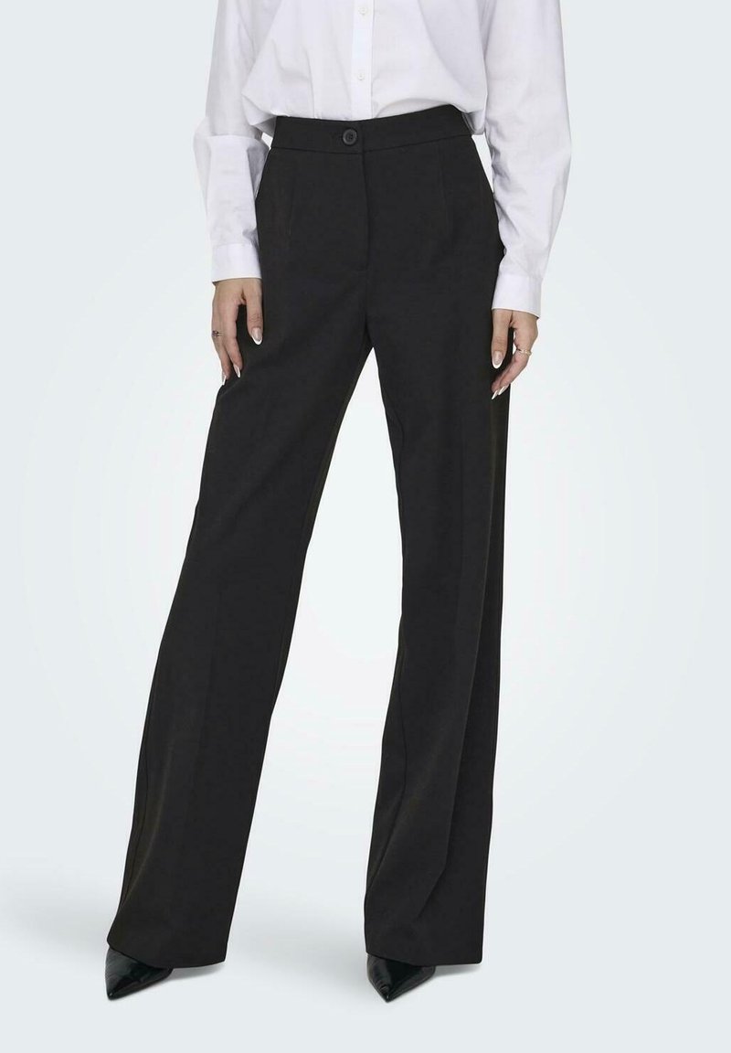 Personne portant un pantalon noir à jambes larges, une chemise blanche à manches longues et des chaussures noires pointues, debout les mains le long du corps.