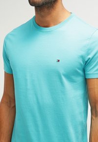 Turquoise cotton t-shirt with a crew neckline and short sleeves. Features a small logo in red, white, and blue on the left chest.
