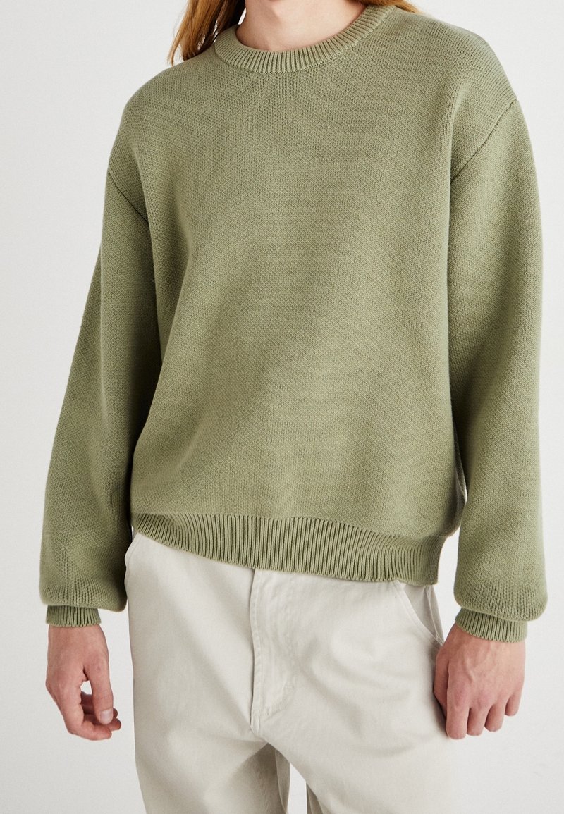 Personne portant un pull en maille vert olive clair avec col, poignets et ourlet côtelés, associé à un pantalon décontracté écru.