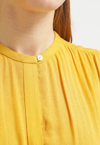 Blusa gialla con scollo rotondo, texture plissettata e un unico bottone visibile al colletto. Tessuto morbido e leggero.