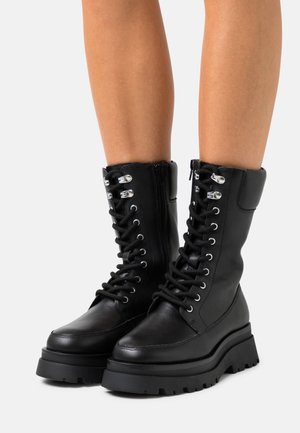 Czarne skórzane buty za kostkę z wiązaniem na sznurówki, o grubych bieżnikowanych podeszwach, noszone na gołych nogach na białym tle.