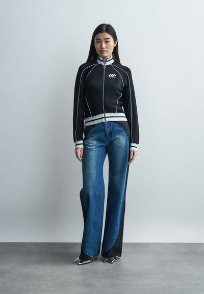 Νεαρή γυναίκα που φοράει μαύρο μπουφάν με φερμουάρ και λευκή ρίγα, καθώς και τζιν παντελόνι με ευρεία άκρα σε δύο τόνους του μπλε, στέκεται μπροστά σε απλό γκρι φόντο.