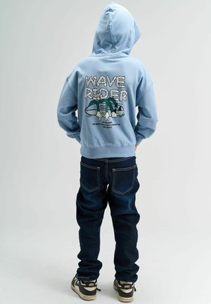 Jasnoniebieska bluza z kapturem z grafiką "WAVE RIDER" na plecach, z ilustracją postaci na desce surferskiej, w połączeniu z ciemnymi dżinsami i sneakersami.