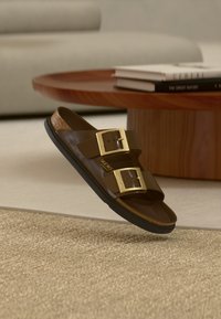 Καφέ σανδάλι με δύο μεγάλες χρυσές αγκράφες αιωρείται πάνω από μια υφασμάτινη μπεζ μοκέτα, κοντά σε έναν στρογγυλό ξύλινο τραπεζάκι με βιβλία.