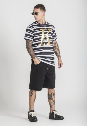 Mies tatuoinneilla, pyöreät aurinkolasit kasvoillaan, mustavalkoraidallinen paita, jossa kultaista "K"-kirjainta, mustat shortsit ja mustat lenkkarit, seisoo kädet taskuissa.