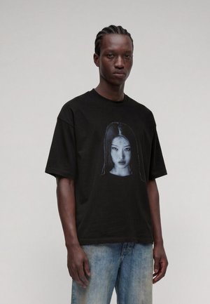 Ung mand med flettet hår iført en sort T-shirt med et sort-hvidt portræt af en kvinde, kombineret med lyseblå jeans.