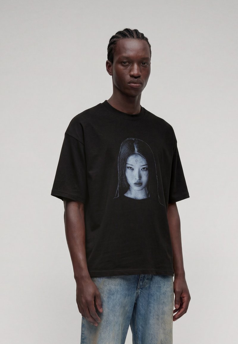 Νέος άνδρας με πλεξούδες που φοράει μαύρο T-shirt με ασπρόμαυρο πορτρέτο γυναίκας, συνδυασμένο με ανοιχτό μπλε τζιν.