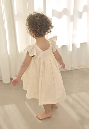 Toddler with curly hair wearing a cream dress, standing barefoot on a smooth floor, with sunlight casting shadows through white curtains.
