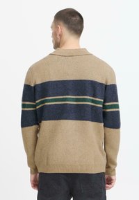 Beige neulepusero, jossa on sinivihreä raidallinen kuvio. Varustettu kauluksella ja ribbireunuksilla, esittelee pehmeää tekstiiliä.