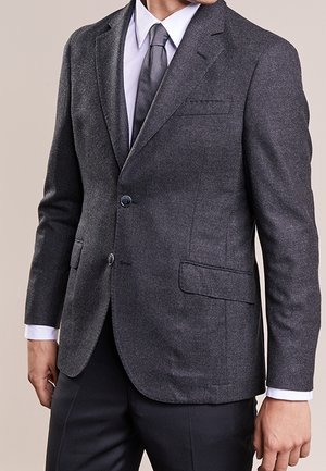 Mann, der einen dunkelgrauen Blazer, ein weißes Hemd und eine graue Krawatte trägt, steht mit entspannten Armen seitlich am Körper vor einem neutralen Hintergrund.