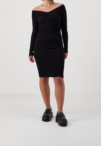 Abito nero a coste con design off-shoulder, maniche lunghe e silhouette aderente, abbinato a mocassini neri con plateau e dettagli in catena.