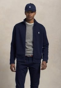Giovane uomo che indossa una giacca a zip blu navy, berretto coordinato con loghi bianchi, maglione grigio, jeans scuri e una cintura marrone, in piedi contro uno sfondo semplice.
