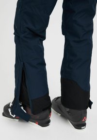 Navy blue ski pants with black accents, featuring side zippers and a fitted design, paired with black ski boots highlighting red details.