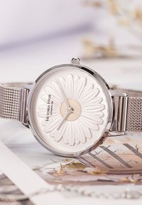 Orologio con cinturino in rete d'argento, caratterizzato da un quadrante bianco con un motivo a margherita in rilievo, cassa circolare e il nome del marchio "Victoria Hyde" in mostra.