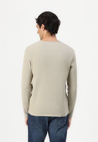 Jersey de manga larga beige claro con un patrón de punto texturizado, escote redondeado y un corte relajado, combinado con jeans de mezclilla oscuros.