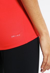 Top deportivo rojo DRI-FIT con una textura suave y elástica y un diseño ajustado, combinado con leggings negros. Detalles de costuras visibles.