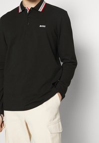 Svart långärmad polo med en röd och vit randig krage, med "BOSS"-logotypen i vitt på bröstet. Kombineras med ljusbeige byxor.