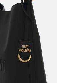 Črna usnjena torbica z embosiranim logotipom in zlatim obeskom z napisom "Love Moschino." Oblika je poenostavljena, material pa mehak.