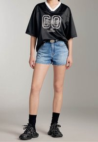 Svart oversize-tröja med vit V-ringning, prydd med en grafisk design. Kombinationen med ljusblå denim-shorts och svarta sportskor.