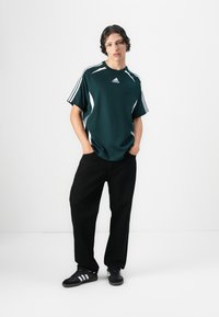 Mørk grønn atletisk skjorte med hvite detaljer, kort erm, ribbet krage, og Adidas-logo. Kombinert med svarte løse bukser og svarte sneakers.
