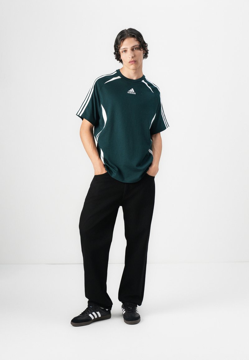 Mørk grønn atletisk skjorte med hvite detaljer, kort erm, ribbet krage, og Adidas-logo. Kombinert med svarte løse bukser og svarte sneakers.