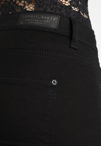 Jeans de mezclilla negra con una textura suave, que presentan una etiqueta "ESPRIT Denim", costuras laterales y un pequeño remache plateado en el bolsillo trasero.