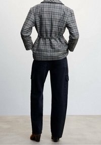 Grijze geruite blazer met een riem in de taille, gecombineerd met donkerblauwe cargobroeken; beide hebben een gestructureerde pasvorm en nette naden. Bruine laarzen met hak.