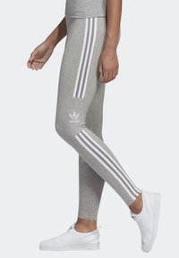 Leggings grigi con tre strisce verticali bianche lungo i lati e un logo Adidas sulla gamba, realizzati in tessuto elasticizzato.