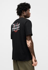 Μαύρο βαμβακερό t-shirt με κοντά μανίκια, με μεγάλο τυπωμένο λογότυπο στην πλάτη σε λευκό και κόκκινο. Μονόχρωμο, άνετη εφαρμογή.