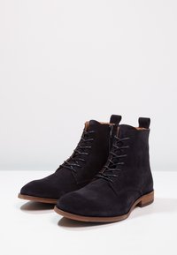 Stivaletti in suede nero alla caviglia con punta arrotondata, chiusura con lacci e sottopiede in legno marrone discreto. Presentano un anello laterale per indossarli facilmente.