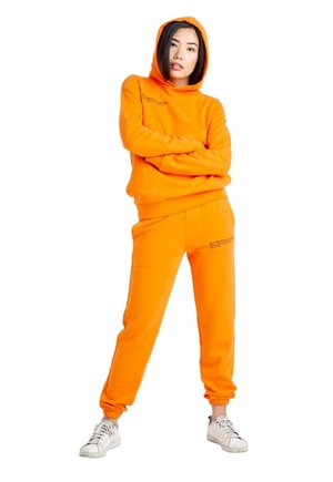 Une femme se tient debout, les bras croisés. Elle porte un sweat à capuche orange vif, un pantalon de jogging assorti et des baskets blanches, sur un fond blanc uni.