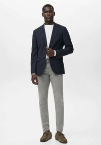 Hombre vistiendo un blazer azul marino, camisa blanca, jeans grises y zapatos de tacón marrones, de pie frente a un fondo blanco liso.