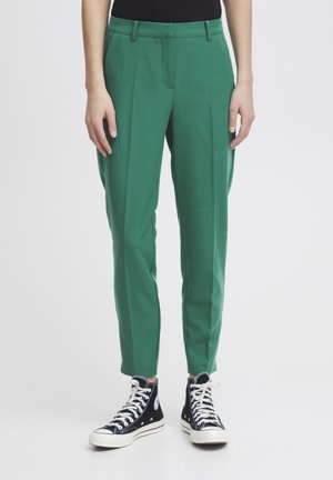 Green tailored trousers with pleats, made from smooth fabric. Features a mid-rise waist and tapered legs, paired with black high-top sneakers.