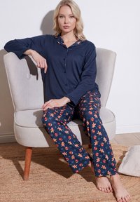Woman wearing navy blue pajama set with gingerbread and heart patterns sits barefoot on light gray armchair.
