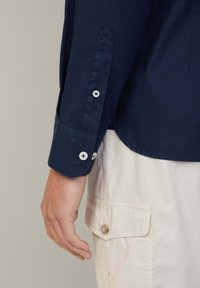 Marineblauwe katoenen shirt met knoopsluiting bij de manchetten en witte knopen; een licht gebogen zoom en opgevouwen mouwdetails; gedragen met lichtgekleurde cargobroek.
