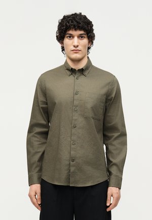 Homme aux cheveux bouclés portant une chemise boutonnée vert olive avec une poche poitrine et un pantalon noir, debout devant un fond clair uni.
