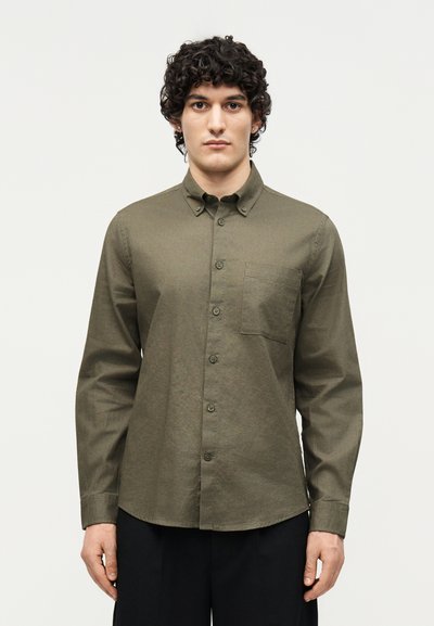 Hombre con pelo rizado que lleva una camisa verde oliva abotonada con bolsillo en el pecho y pantalones negros, de pie frente a un fondo claro y liso.