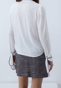 Biała bluzka z falbanianym kołnierzem i długimi rękawami, z czarnymi akcentami przy otworach rękawów, w połączeniu z teksturowaną, ciemną wzorzystą spódnicą.