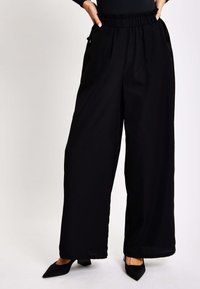 Black wide-leg trousers with an elastic waistband and side pockets; smooth texture and flowy silhouette; paired with black pointed-toe heels.