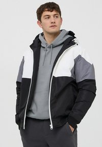 Giacca imbottita nera, bianca e grigia con zip frontale, cappuccio e design a blocchi di colore. Indossata sopra un hoodie grigio con lacci.