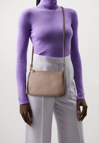 Beige læder crossbody taske med tekstureret finish, buet form, guldfarvet hardware og justerbar skulderrem, båret over lysegrå bukser.