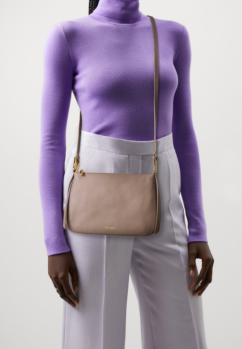 Beige læder crossbody taske med tekstureret finish, buet form, guldfarvet hardware og justerbar skulderrem, båret over lysegrå bukser.