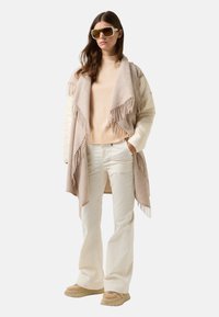 Vrouw in oversized beige zonnebril, crème turtleneck, franjesjaal, gewatteerde jas, witte broek en beige sneakers, staand met handen in de zakken.