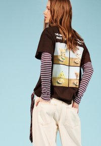 Braunes Grafik-T-Shirt mit Cartoon-Bären und einem Küken auf einem hellen Hintergrund, über einem gestreiften Langarmshirt getragen.