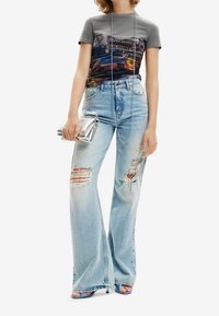 Grå grafisk t-shirt med et farverigt print, parret med lyseblå vide bukser med slid og huller. Metalisk clutchtaske og stilige hæle.