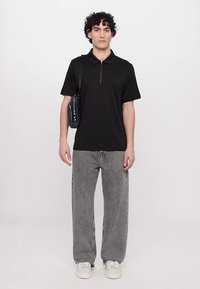 Svart polo-skjorte med korte ermer og glidelås krage, sammen med løse grå jeans og hvite sneakers. Modellen holder en marineblå veske.