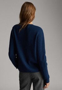 Ciemnoniebieski sweter prążkowany z okrągłym dekoltem i długimi rękawami, zestawiony z szarymi spodniami, widziane z tyłu.